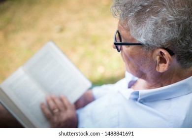 Profile Portrait Of An Older Caucasian Man Reading A Book With Glasses In Outdoor Location.