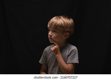 Profile portrait of pensive handsome blonde little boy in gray T-shirt with finger near mouth on black background. Mute boy speaks with gestures. Counts in mind or remembers poem. Kid think or compos.
