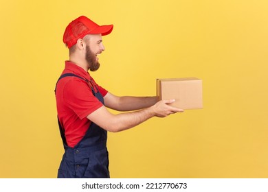 Profile Portrait Of Positive Deliveryman Holding Cardboard Parcel, Delivering Order Door-to-door, Shipment And Cargo Transportation Service. Indoor Studio Shot Isolated On Yellow Background.