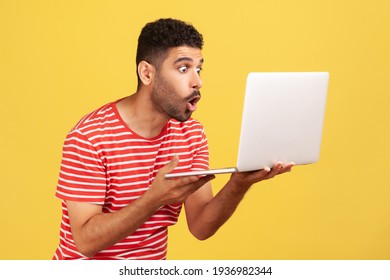 Profile Portrait Shocked Surprised Man With Beard In Striped T-shirt Holding Laptop And Looking At Display With Big Astonished Eyes. Indoor Studio Shot Isolated On Yellow Background