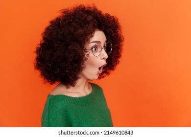 Profile Portrait Of Shocked Woman With Afro Hairstyle In Green Casual Style Sweater Standing With Open Mouth, Looking Away, Sees Something Surprising. Indoor Studio Shot Isolated On Orange Background.