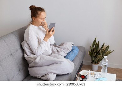 Profile portrait of sick female wearing white sweater sitting on sofa wrapped in blanket, holding phone in hands, checking social networks while staying home during virus illness.