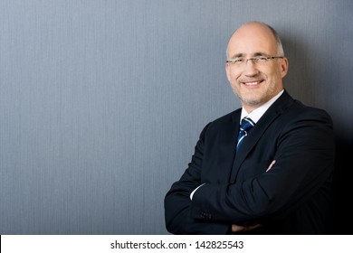 Profile Portrait Of A Smiling And Confident Mature Balding Businessman, Wearing A Suit And A Necktie, With Arms Crossed, In Front Of A Gray Wall With Copy-space