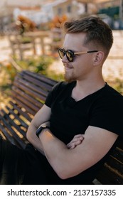 Profile portrait of stylish handsome man with blond hair wearing black t-shirt and sunglasses siting on the bench with ocean view in sunny warm day. High quality photo
