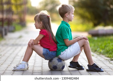 Profile Portrait Of Two Cute Blond Children, Smiling Boy And Long-haired Girl Sitting On Soccer Ball On Empty Sunny Suburb Paved Road Blurred Summer Bright Background. Children Games And Fun Concept.