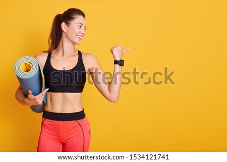Profile portrait of young woman holding mat and shows muscles, sporty female looking aside, wearing stylish sportwear, poses smiling in studio isolatedover yellow background. Healthy lifestyle concept