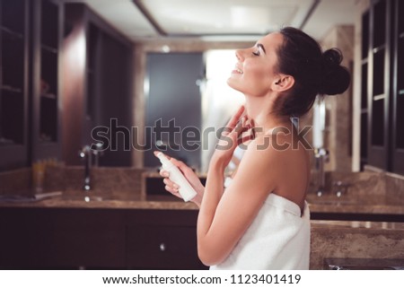 Profile of satisfied woman applying cream on neck with joy. She is holding cosmetic container in one hand while gently touching skin with another. Copy space in left side