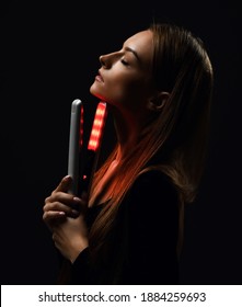 Profile of sensual young woman with silky straight hair standing with her eyes closed holding white modern hair straightener at face chin over dark background. Haircare, beauty, wellness concept
