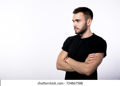 in profile serious young brown-haired man with a small stylish beard and mustache in a black T-shirt crossed his arms over his chest against a white background