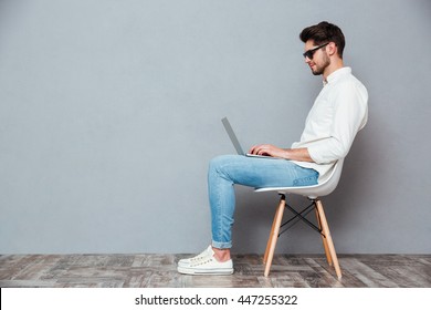 Profile Of Serious Young Man In Sunglasses Sitting On Chair And Using Laptop Over Grey Background