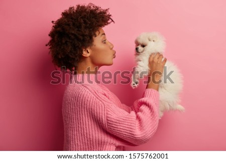 Profile shot of Afro American woman kisses breed dog, raises in hands, wears knitted sweater, poses against pink background, has playful mood, expresses love, likes playing with small puppy.