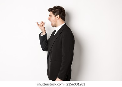 Profile Shot Of Angry Businessman In Black Suit, Shouting At Speakerphone And Looking Mad, Recording Voice Message, Standing Over White Background