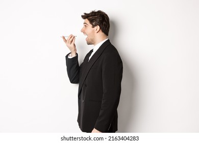 Profile Shot Of Handsome Businessman In Black Suit Talking On Speakerphone, Smiling And Looking Happy, Recording Voice Message, Standing Over White Background