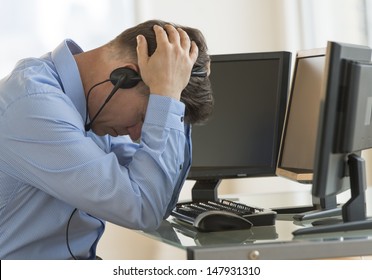 Profile shot of male exhausted trader with head in hands leaning at computer desk in office