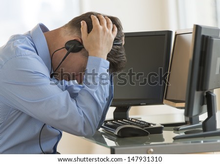 Profile shot of male exhausted trader with head in hands leaning at computer desk in office