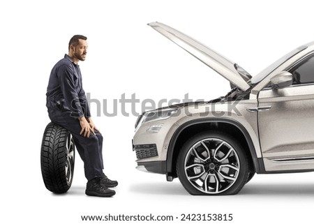 Profile shot of a mechanic sitting on a car tire and looking at a SUV with open hood isolated on white background