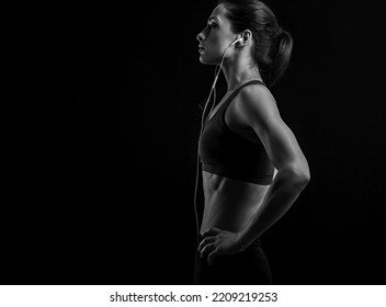 Profile Shot Of Serious Female Athlete With Ponytail, Listening Music In Headphones In Sportsbra, Standing Isolated On Black Background With Empty Copy Space. Lifestyle Portrait. Black And White