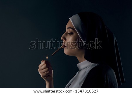 Profile shot of upper nun's body, posing on black background. She wearing dark nun's clothing. The nun is lighting a cigarette. She looking down. 