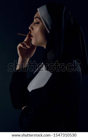 Profile shot of upper nun's body, posing on black background. She wearing dark nun's clothing. The nun is taking a draw at a cigarette. Her eyes are close. 