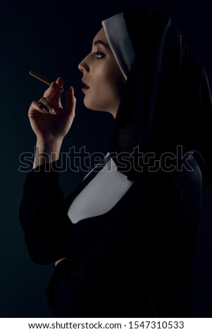 Profile shot of upper nun's body, posing on black background. She wearing dark nun's clothing. The nun is smoking and looking forward. 
