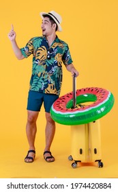 Profile Shot, Young Asian Man In Colorful Hawaiian Shirt Gives Index Finger Up And Pull Yellow Suitcase. Full Body Studio Portrait On Yellow Background. Happy Summer Holiday Travel Concept