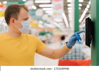 Profile Shot Of Young Man Types On Touch Screen Of Self Checkout, Wears Protective Rubber Blue Gloves, Poses Against Blurred Supermarket Background. Consumerism, Shopping, Quarantine Concept