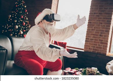 Profile side photo addicted grey beard santa claus sit couch play 3d simulation game on x-mas christmas party use modern goggles lose wear red cap headwear in house indoors evergreen tree - Powered by Shutterstock