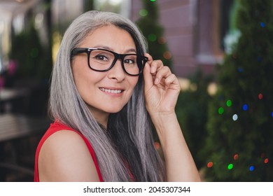 Profile side photo of mature attractive asian woman happy positive smile hands touch glasses glad outdoors