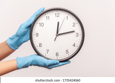 Profile Side View Closeup Of Human Hand In Blue Surgical Gloves Holding Analog Clock. Indoor, Studio Shot, Isolated On Gray Background.