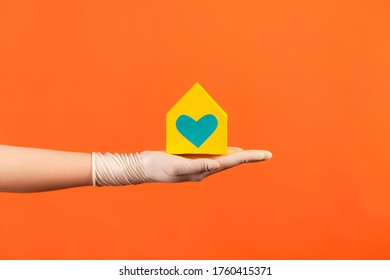 Profile Side View Closeup Of Human Hand In White Surgical Gloves Holding Yellow Paper House Exterior In Hand With Love And Care. Indoor, Studio Shot, Isolated On Orange Background.