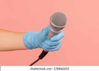 Profile side view closeup of human hand in blue surgical gloves holding silver microphone. indoor, studio shot, isolated on pink background.