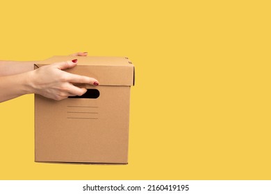 Profile Side View Closeup Of Human Hand Holding Cardboard Box, Carton Parcel, Delivery Concept. Indoor Studio Shot Isolated On Yellow Background.