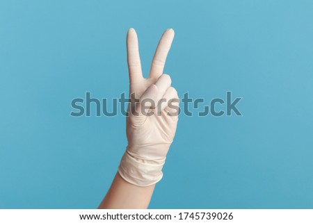 Profile side view closeup of human hand in white surgical gloves showing victory, peace sign or number 2 with fingers. indoor, studio shot, isolated on blue background.