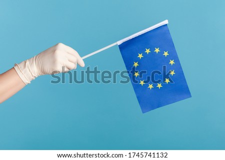 Profile side view closeup of human hand in white surgical gloves holding Eroupean union flag. indoor, studio shot, isolated on blue background.