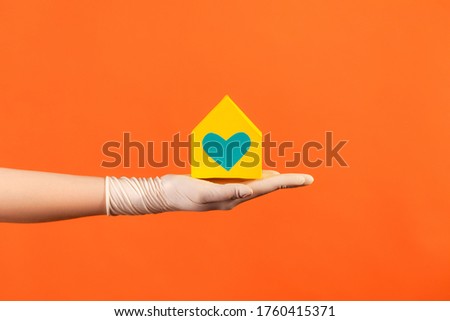 Profile side view closeup of human hand in white surgical gloves holding yellow paper house exterior in hand with love and care. indoor, studio shot, isolated on orange background.