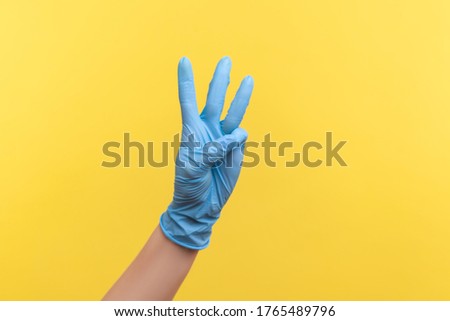 Profile side view closeup of human hand in blue surgical gloves showing number 3 three with hands. indoor, studio shot, isolated on yellow background.