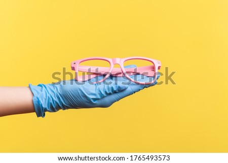 Profile side view closeup of human hand in blue surgical gloves holding and giving pink eyeglasses frame. indoor, studio shot, isolated on yellow background.