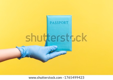 Profile side view closeup of human hand in blue surgical gloves holding passport in hand. indoor, studio shot, isolated on yellow background.