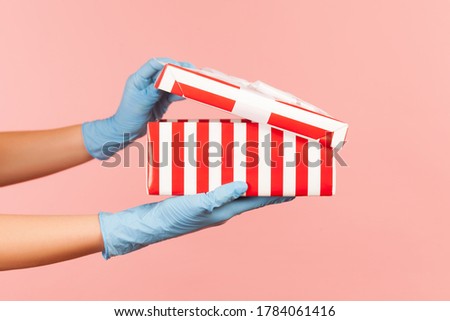 Profile side view closeup of human hand in blue surgical gloves holding and opening striped red white gift box. indoor, studio shot, isolated on pink background.