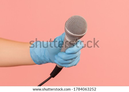Profile side view closeup of human hand in blue surgical gloves holding silver microphone. indoor, studio shot, isolated on pink background.