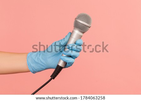 Profile side view closeup of human hand in blue surgical gloves holding silver microphone. indoor, studio shot, isolated on pink background.