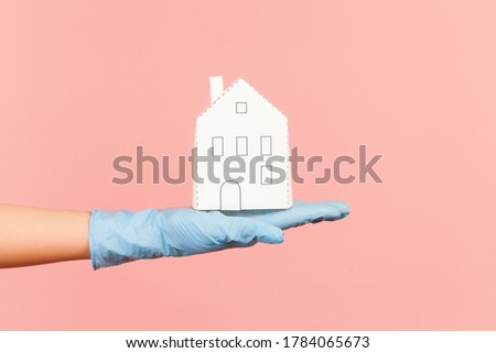 Profile side view closeup of human hand in blue surgical gloves holding white paper house exterior in hand. indoor, studio shot, isolated on pink background.