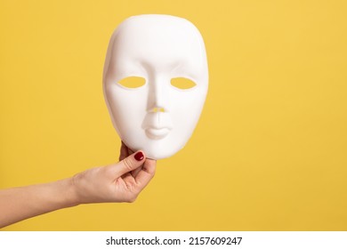 Profile side view closeup of woman hand holding white facial mask model, hiding personality. Indoor studio shot isolated on yellow background.