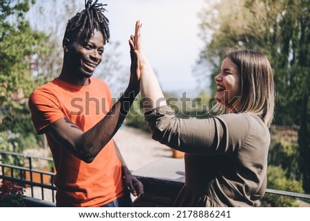 Profile side view of happy african man making greeting gesture give high five with smiling and young caucasian woman. Two best friends clapping hands, standing outside in the park - friendship concept