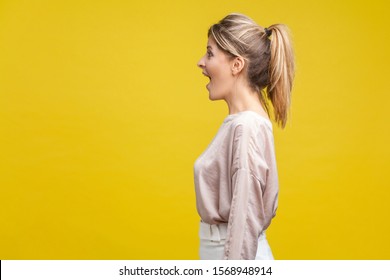 Profile side view of happy amazed blonde woman with ponytale hairstyle and in casual beige blouse standing, looking with surprised face, open mouth, indoor studio shot isolated on yellow background