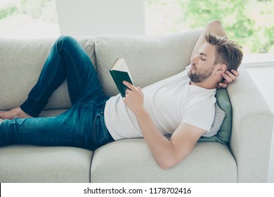 Profile side view photo of attractive, bearded, good-looking man in white t-shirt lying in bed, reader book after hard working week in light modern interior