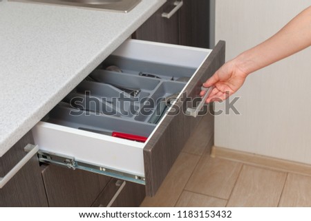 Profile side view photo of part clean white cook table, woman hand open new brown kitchen drawer by modern door handle, with different cutlery spoon, pizza knife, fork and stuff