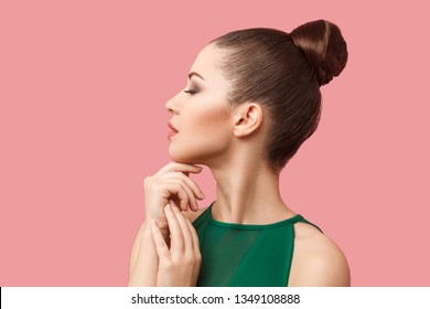 Profile Side View Portrait Of Calm Serious Beautiful Young Woman With Bun Hairstyle And Makeup In Green Dress Standing With Closed Eyes And Touching Her Chin. Studio Shot, Isolated On Pink Background.