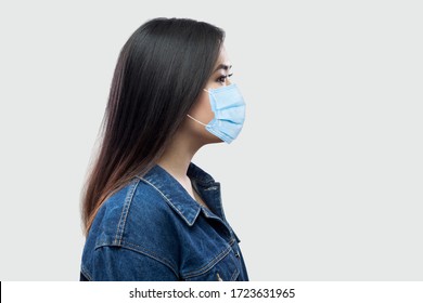 Profile Side View Portrait Of Calm Serious Beautiful Brunette Asian Young Woman With Surgical Medical Mask In Blue Jacket Standing And Looking. Indoor Studio Shot, Isolated On Light Grey Background.