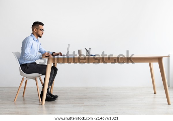 Profile Side View Portrait Of Focused Middle\
Eastern Male Manager In Glasses Working On Laptop Computer In\
Modern Office, Serious Guy Sitting At Desk And Using Pc, Looking At\
Screen Watching Webinar